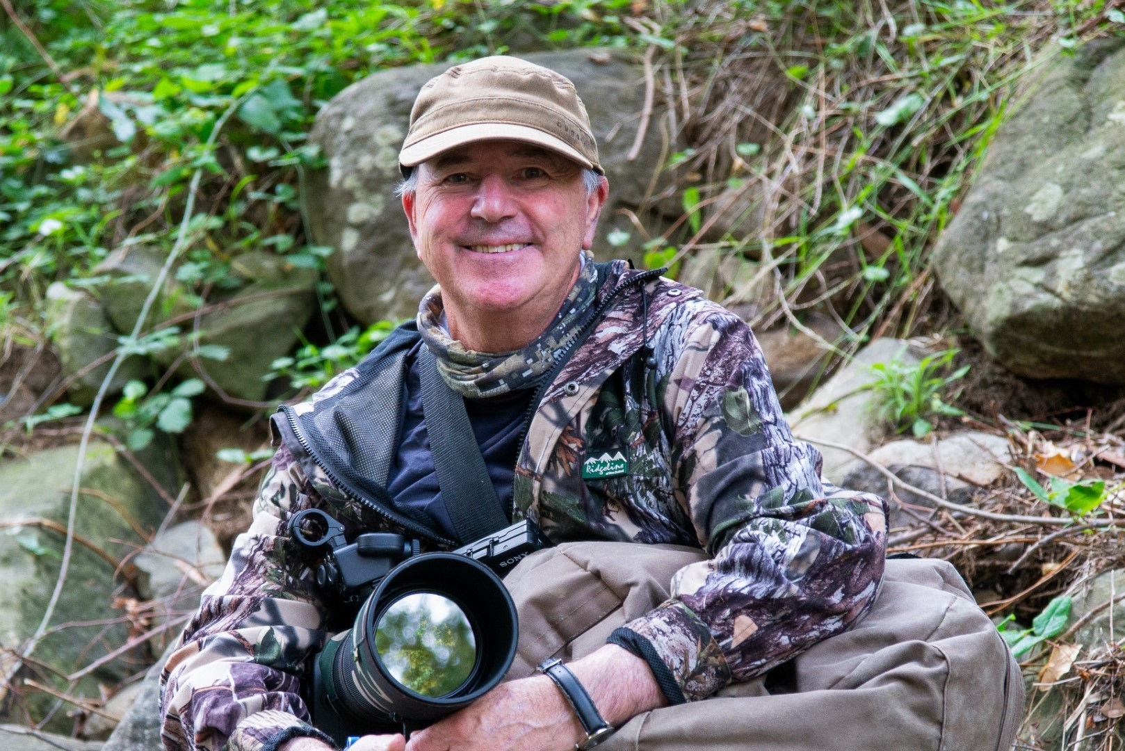 man in camouflage sitting on a rock with a large camera 